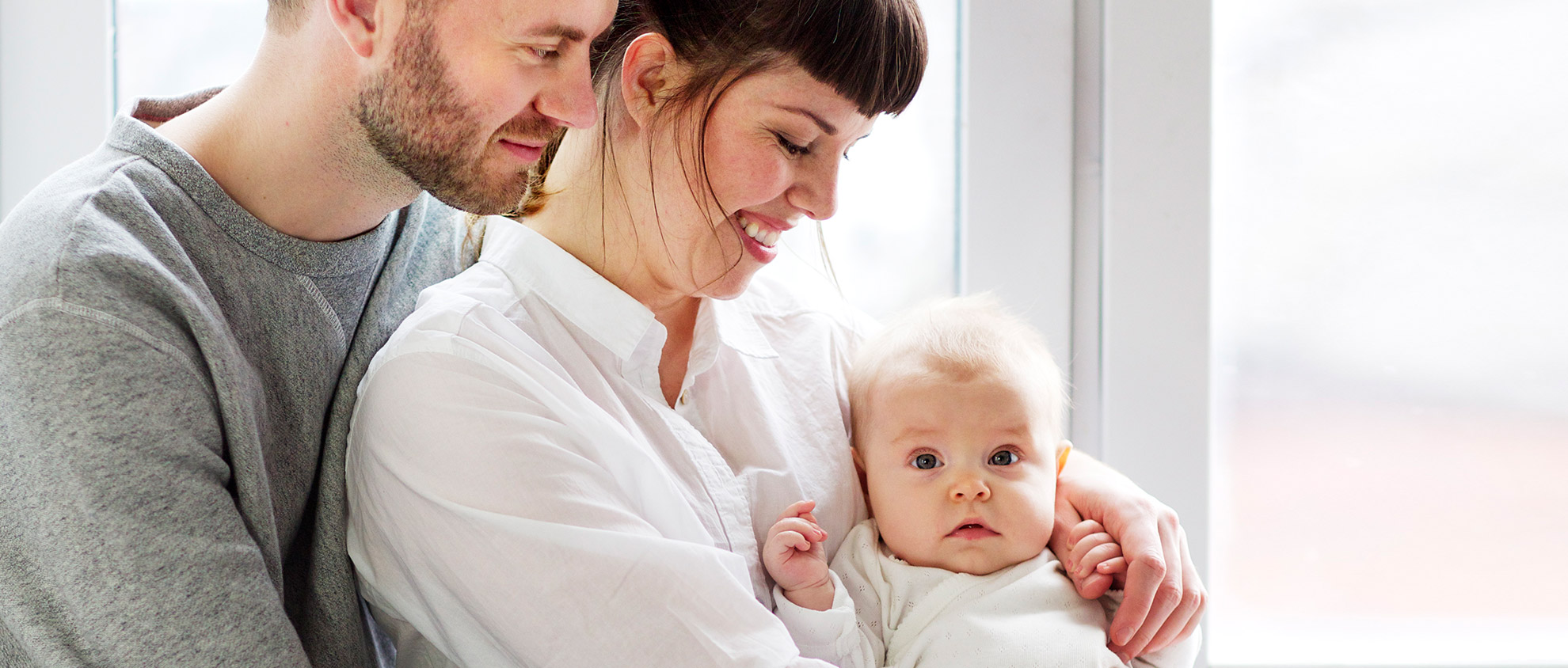 En man och en kvinna, klädda i fritidskläder, håller en bebis tätt intill sig och tittar kärleksfullt på barnet.