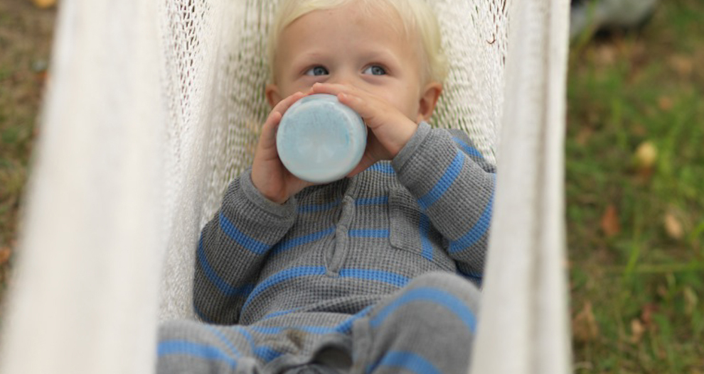 Ett litet barn i randig klädsel sitter i en hängmatta och dricker ur en flaska.