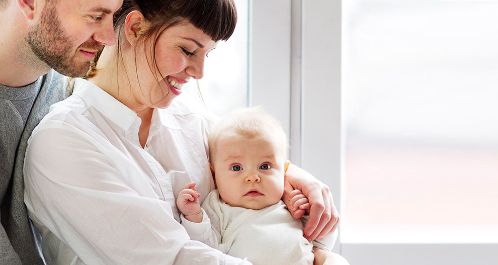 En man och en kvinna, klädda i fritidskläder, håller en bebis tätt intill sig och tittar kärleksfullt på barnet.