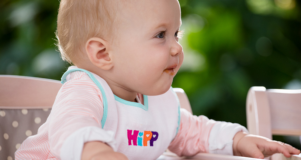 Ett barn i en barnstol, med en haklapp med ordet "HIPP", tittar åt sidan.