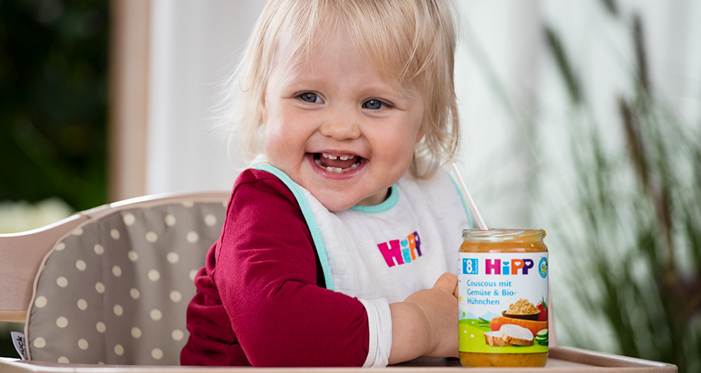 Ett litet barn sitter i en barnstol, iklädd haklapp och röd tröja, med en burk med mat bredvid sig.